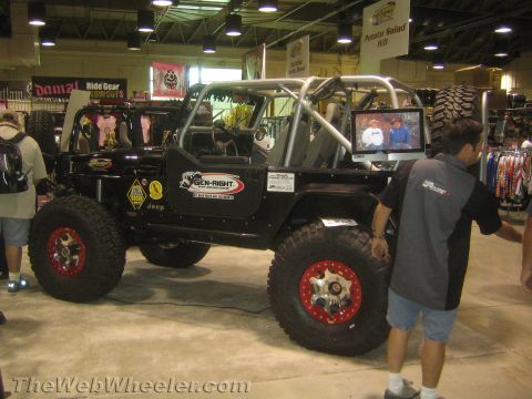1990 Ford Bronco - black lifted Jeep with a roll cage, oversized mud tires on red beadlock wheels and Genright sponsor graphics on display at an expo.