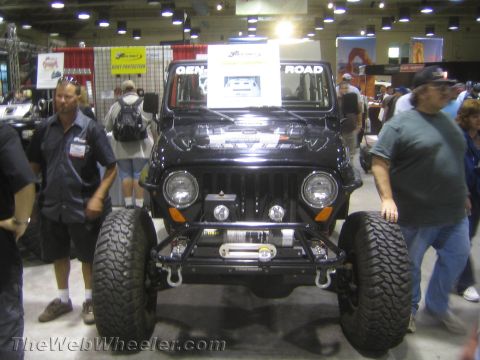 1990 Ford Bronco - black lifted Jeep Wrangler with a front winch bumper and large mud tires sits on display amid crowds at an indoor off-road trade show.