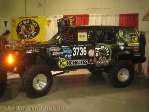 1990 Ford Bronco - olive green Jeep race truck marked 3736 with KC Hilites and sponsor decals sits before a Pit Bull Tires banner at an indoor booth.