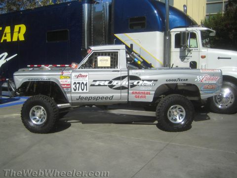 1990 Ford Bronco - silver Jeepspeed race truck numbered 3701 with sponsor decals sits on pavement next to a dark transport semi-trailer.