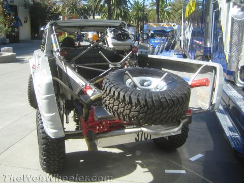 1990 Ford Bronco - rear of a stripped tube-chassis race truck shows its mounted spare tire, jack and roll cage hardware on outdoor pavement.