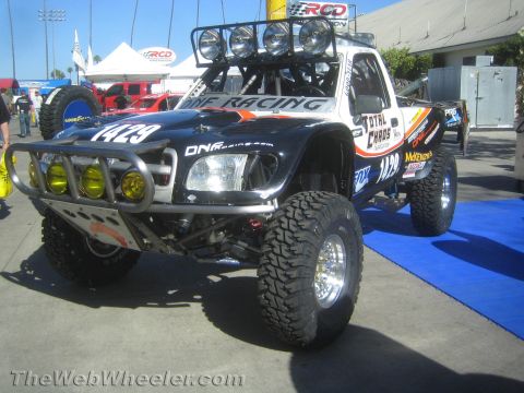 1990 Ford Bronco - black off-road prerunner race truck marked 1429 with a roof light bar and yellow auxiliary lights sits on a blue carpet at an outdoor expo.