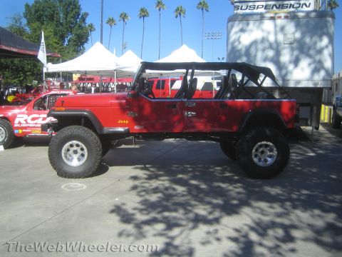 1990 Ford Bronco - red stretched multi-door Jeep with an extended roll cage and large tires is shown in profile near vendor tents at an outdoor event.
