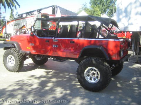 1990 Ford Bronco - red stretched multi-door Jeep with exposed roll cage seating sits on a suspension lift and large tires at an outdoor display.