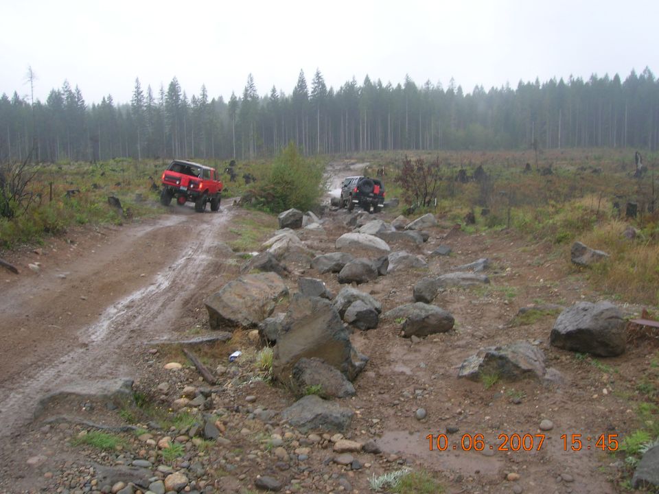 Two off-road vehicles navigate a muddy, rocky trail through a deforested area with a line of trees in the background