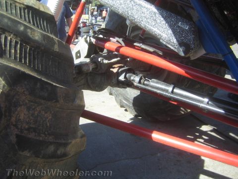 1990 Ford Bronco - close-up shows red-painted suspension links, a tie-rod and driveshaft components next to a muddy off-road tire.