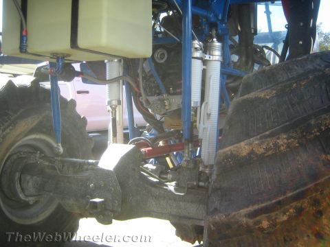1990 Ford Bronco - close-up of a rear suspension shows blue control arms, a coilover shock and reservoir beside a large mud tire.
