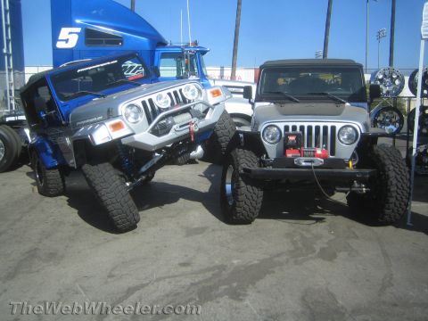 1990 Ford Bronco - blue and a silver lifted Jeep Wrangler with front winches and large tires sit side by side on pavement at an outdoor event.