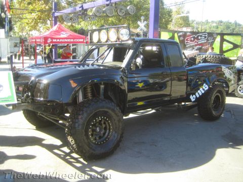 1990 Ford Bronco - black extended-cab prerunner pickup with a roof light bar and BMR decals sits on display among vendor tents outdoors.