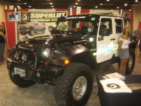 1990 Ford Bronco - black Jeep Wrangler Rubicon with auxiliary lights and a winch sits at a Superlift suspension display booth at an indoor expo.