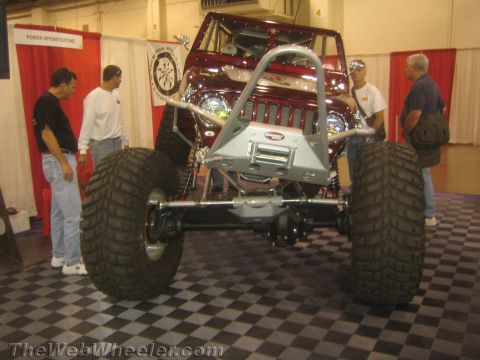 1990 Ford Bronco - dark red tube-chassis rock crawler with oversized tires and a silver winch bumper sits on display at an indoor trade show as spectators loo…