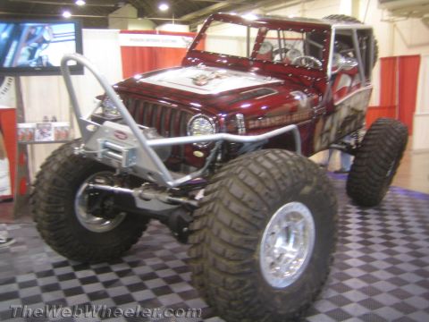 1990 Ford Bronco - maroon tube-chassis rock buggy with airbrushed graphics, a silver winch bumper and big mud tires sits on display under show lighting.