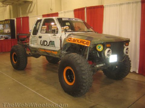 1990 Ford Bronco - camouflage-and-white lifted pickup rock crawler on orange beadlock wheels and big tires sits on display at an indoor expo.
