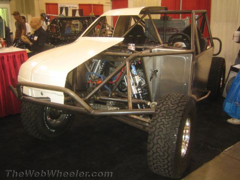 1990 Ford Bronco - unpainted tube-chassis buggy with a white fiberglass nose, exposed engine and roll cage sits on display at an indoor expo.