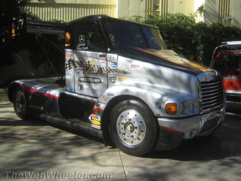 1990 Ford Bronco - silver-wrapped Freightliner cabover-style racing semi tractor with Mercedes-Benz power and Michelin branding parked outdoors among trees.
