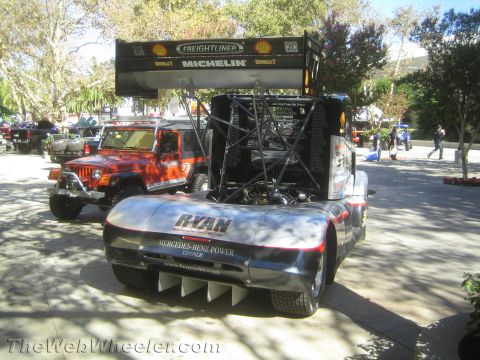 1990 Ford Bronco - rear of a silver Freightliner racing semi with a large roof wing and exposed engine sits outdoors with a red Jeep parked behind.