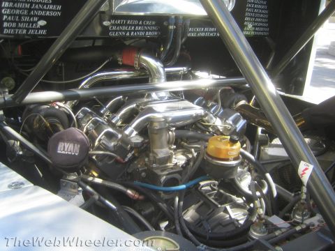 1990 Ford Bronco - polished V8 engine with chrome plumbing sits in a tube-chassis race vehicle below painted sponsor and crew names on the bodywork.
