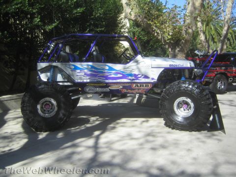 1990 Ford Bronco - white-and-blue flamed tube-chassis Jeep-bodied rock crawler on big tires and blue shocks sits on a paved outdoor display.