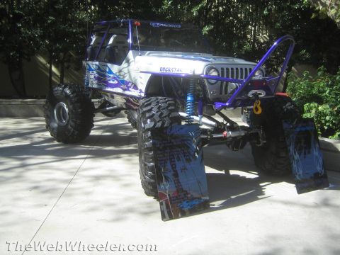 1990 Ford Bronco - white-and-blue tube-chassis rock crawler with a winch and printed ramp boards sits front-on at a paved outdoor display.