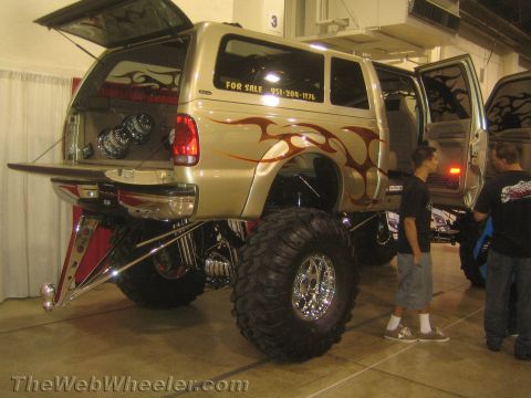 1990 Ford Bronco - tan flame-painted lifted SUV with a chrome rear tube bumper and For Sale sign sits at an indoor show as onlookers inspect it.
