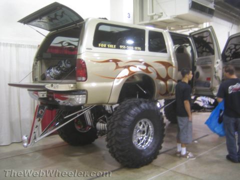 1990 Ford Bronco - tan flame-painted lifted SUV on chrome wheels and mud tires with a For Sale sign sits at an indoor show with spectators beside it.