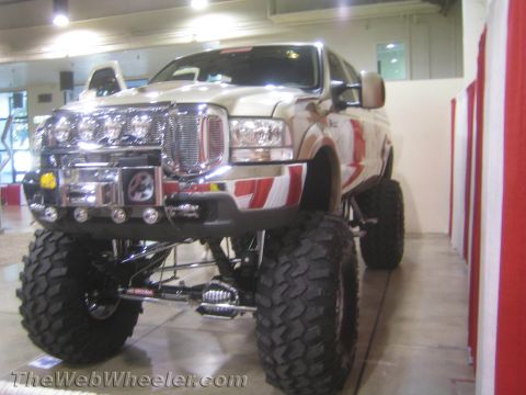 1990 Ford Bronco - tan lifted full-size SUV with a roof light bar, multiple driving lights and a winch bumper sits on display in an indoor hall.