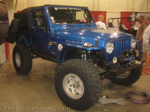 1990 Ford Bronco - blue Jeep Wrangler Rubicon with a winch bumper and oversized mud tires displayed at an indoor expo booth.