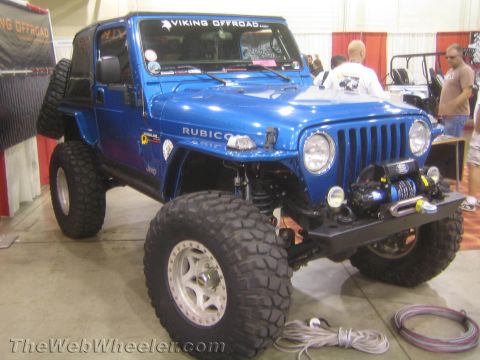 1990 Ford Bronco - lifted blue Jeep Wrangler Rubicon with a front winch and big mud tires shown at an indoor off-road show.