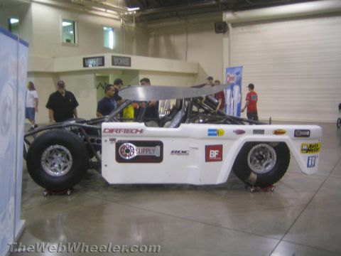 1990 Ford Bronco - Side profile of a white-bodied tube-chassis off-road race car with a roof wing, parked on dollies at a show.