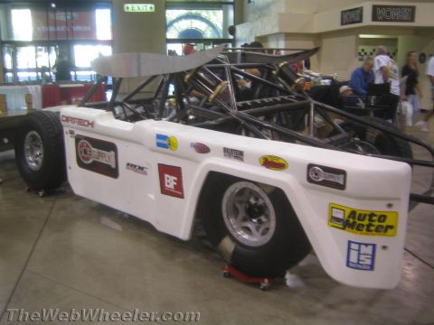 1990 Ford Bronco - Rear three-quarter view of a white tube-chassis off-road race car covered in sponsor decals, indoors on dollies.