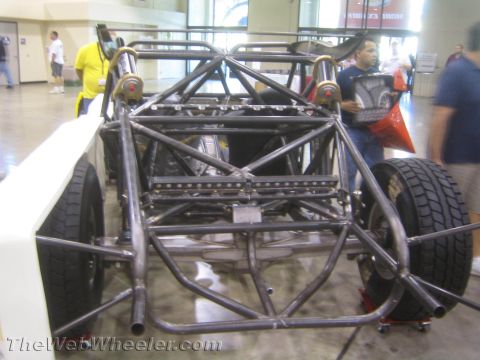 1990 Ford Bronco - rear of a bare welded tube-frame off-road race chassis with exhaust headers and a rear tire, shown indoors.