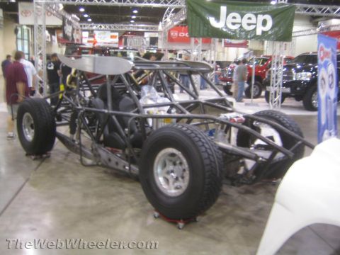 1990 Ford Bronco - bare tube-chassis off-road race car sits on dollies at an expo beneath a green Jeep banner.