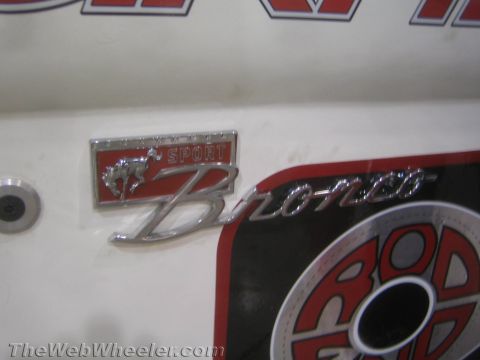 1990 Ford Bronco - chrome Bronco Sport emblem with a running-horse motif mounted on a white body panel beside red graphics.