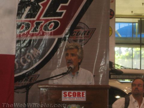 1990 Ford Bronco - gray-haired man with a mustache speaks at a SCORE podium in front of a racing radio banner.