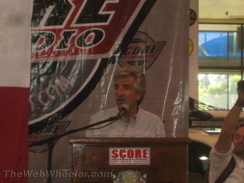 1990 Ford Bronco - man speaks into a microphone at a SCORE off-road racing podium with a radio banner behind him.