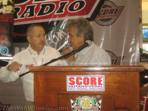 1990 Ford Bronco - Two men stand at a SCORE off-road racing podium under a racing radio banner, one speaking into the mic.