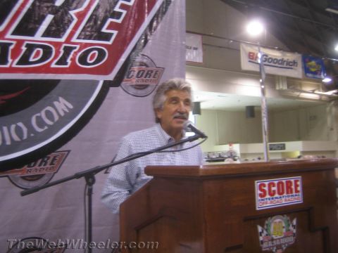 1990 Ford Bronco - gray-haired man speaks at a SCORE podium with racing radio and BFGoodrich banners in the background.
