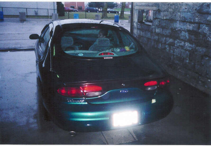 Rear view of dark green Ford Taurus sedan by a brick wall, Flowmaster sticker in rear window, plate obscured by flash