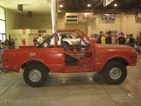1990 Ford Bronco - weathered red vintage pickup-based off-road racer with an exposed interior roll cage sits on display at an indoor automotive show.