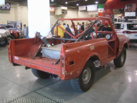 1990 Ford Bronco - rear of a faded red off-road race truck reveals its open bed, roll cage and chunky tires at an indoor exhibition.