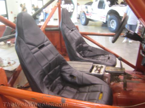 1990 Ford Bronco - pair of padded black bucket race seats mounted inside the red roll cage of an off-road vehicle on display indoors.