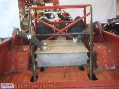 1990 Ford Bronco - Looking into the bed of a red off-road truck shows a metal fuel tank, plumbing and X-braced roll cage tubing.