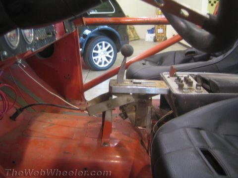 1990 Ford Bronco - floor shifter rises between the worn metal tunnel and a black seat inside an off-road truck, with a blue car visible through the cage.