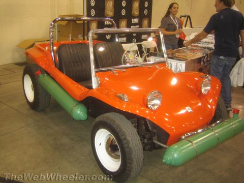 1990 Ford Bronco - glossy orange fiberglass dune buggy with green side fuel cylinders and white wheels sits on display as people browse a vendor booth behind…