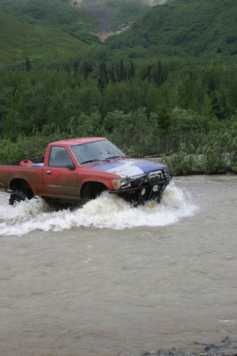 2003 Jeep TJ - A red pickup truck navigates through a shallow stream in a wooded, mountainous area.
