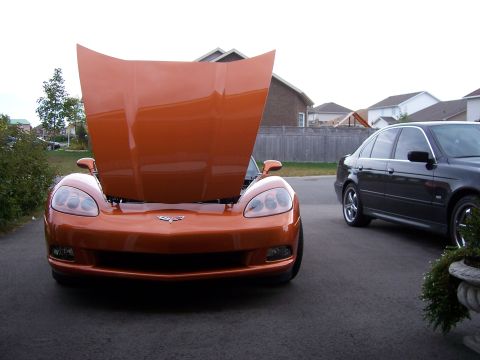 1996 Ford Bronco - An orange Chevrolet Corvette with its hood open is parked in a driveway next to a dark sedan.