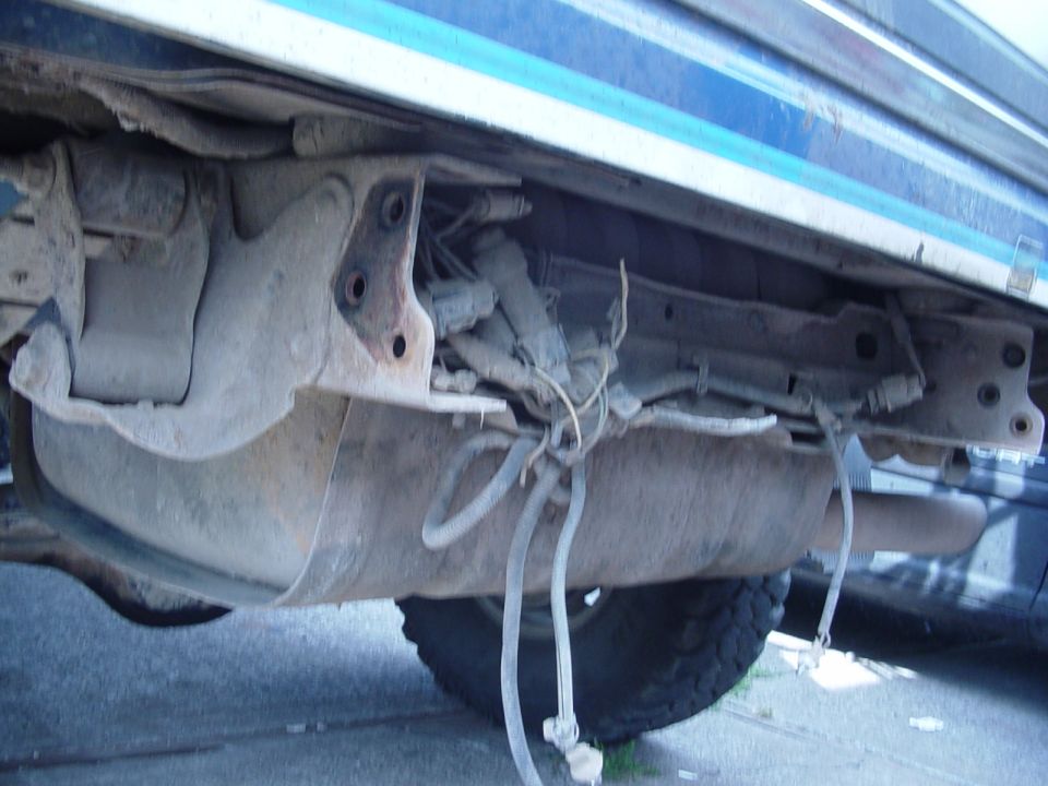 Underbody view showing fuel tank, exhaust pipe, and suspension components. Tire visible