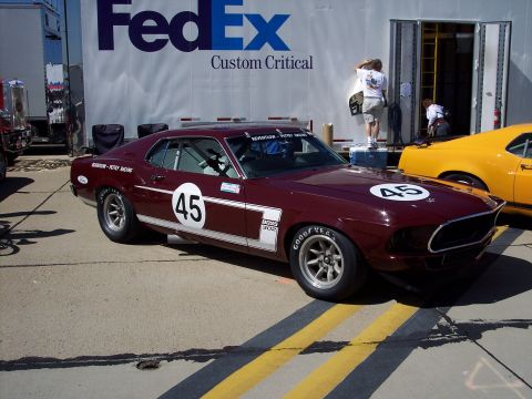 1971 Ford Mustang - A vintage Ford Mustang race car with racing decals and numbers is parked on a track next to another car, with a FedEx truck behind it.