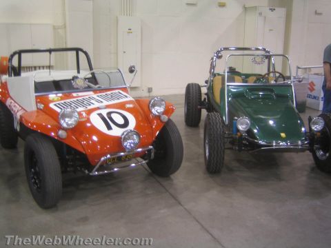 1990 Ford Bronco - Two vintage Manx-style dune buggies sit side by side on a show floor, an orange number 10 racer on the left and a green buggy on the right.