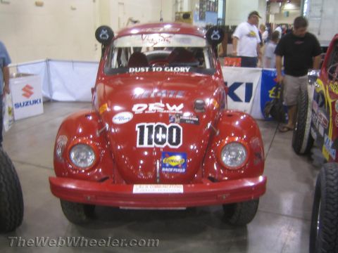 1990 Ford Bronco - red Baja Bug VW Beetle race car wearing number 1100 and Dust to Glory and Tecate livery sits on an indoor show floor flanked by other race…